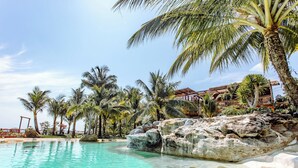 Una piscina turquesa y transparente rodeada de palmeras y formaciones rocosas, con un edificio de techo de paja al fondo.