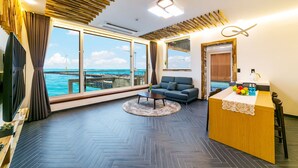 Una habitación moderna con suelo de espiga, un sofá, una mesa de centro y un escritorio con un jarrón de frutas. Amplias ventanas ofrecen vistas al océano y a aerogeneradores.