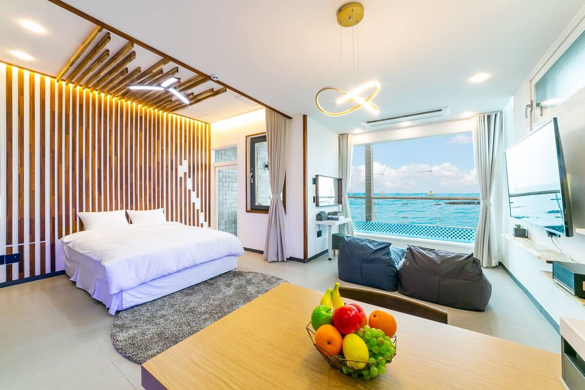 Une chambre d’hôtel moderne avec un lit, une table à manger avec des fruits, une télévision et une vue sur la mer.
