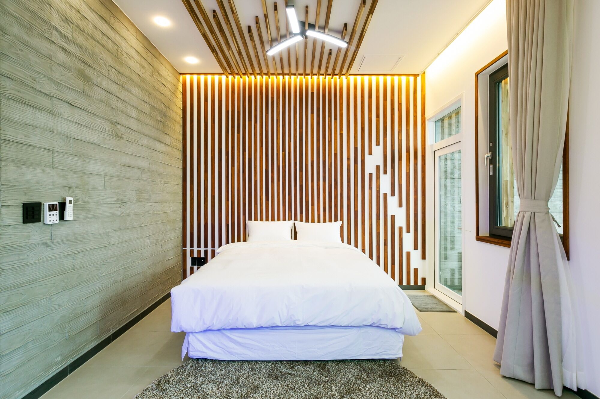 A modern bedroom with a large bed, wooden panel wall, ceiling lights, and a window with curtains.