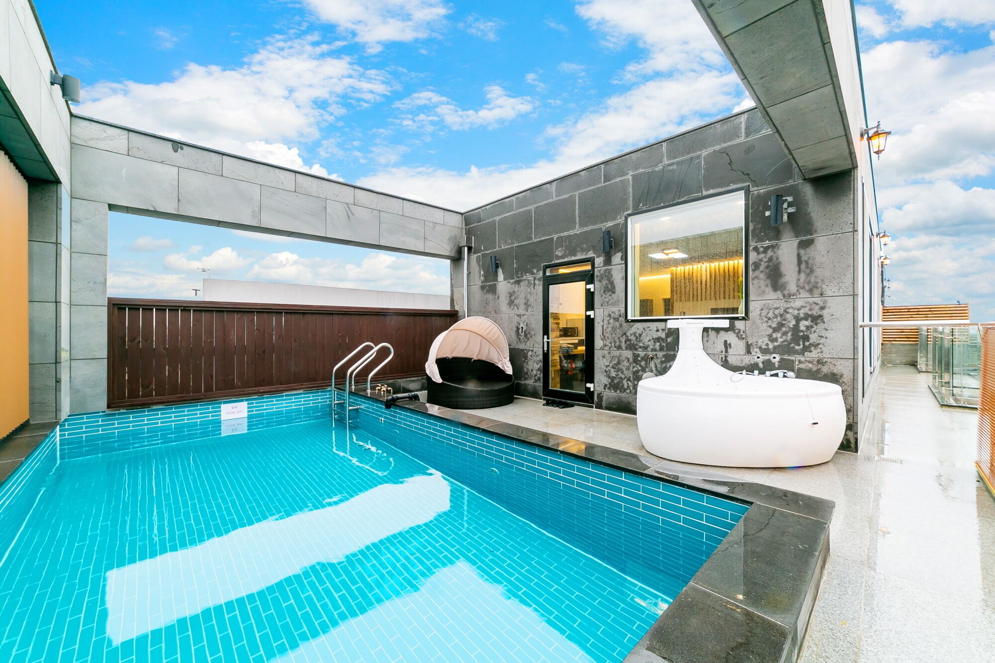 Un área de piscina exterior moderna con una piscina azul cristalina, una bañera blanca grande y una terraza de madera.