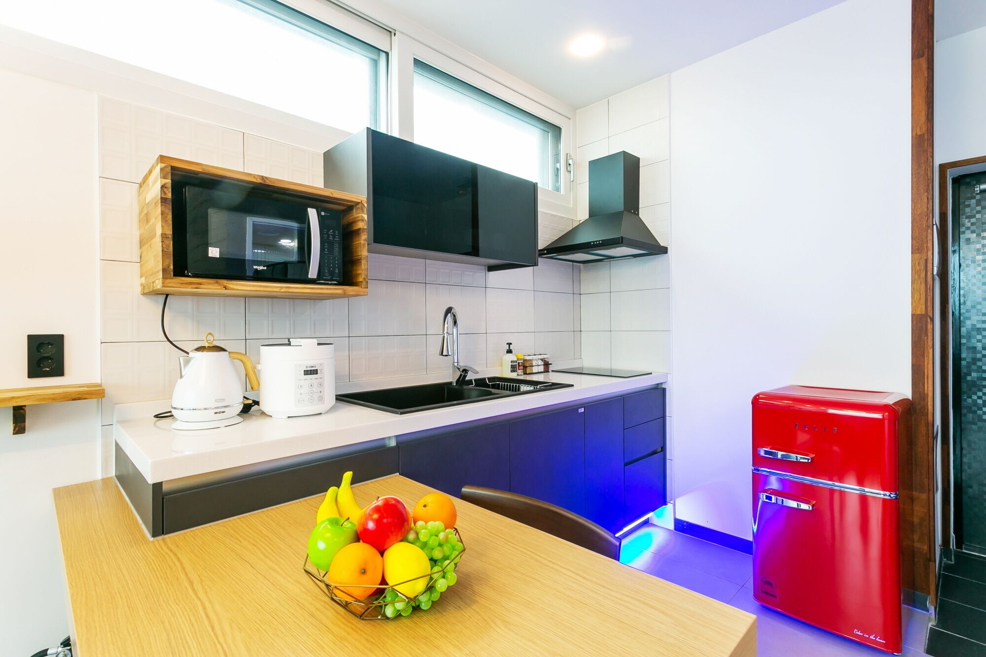 A modern kitchen with white tiles, a microwave, a sink, a red refrigerator, and a fruit bowl on the counter.