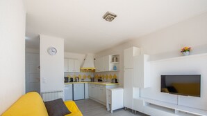 Een moderne woonkamer met een gele bank, een flatscreen televisie aan de muur en een keuken met witte kastjes en gele tegels.