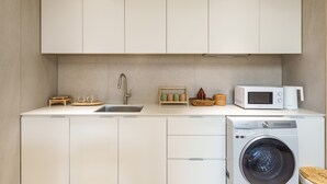 Une cuisine compacte équipée d’un évier, d’un four à micro-ondes, d’un lave-linge Samsung et d’une petite étagère en bois où sont posées des tasses et un panier.