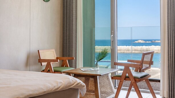 Une chambre d’hôtel avec un lit, deux chaises en bois avec des coussins, une petite table avec une plante et une vue sur l’océan par une porte vitrée.