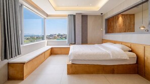 Une chambre d’hôtel moderne dotée d’une grande fenêtre, d’une banquette intégrée, d’un lit et d’une tête de lit en bois.