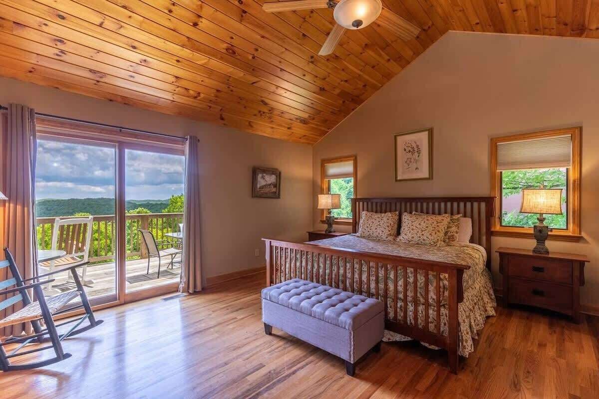 A bedroom with a wooden ceiling, a large bed, a bench, a nightstand, and a view of the outdoors.