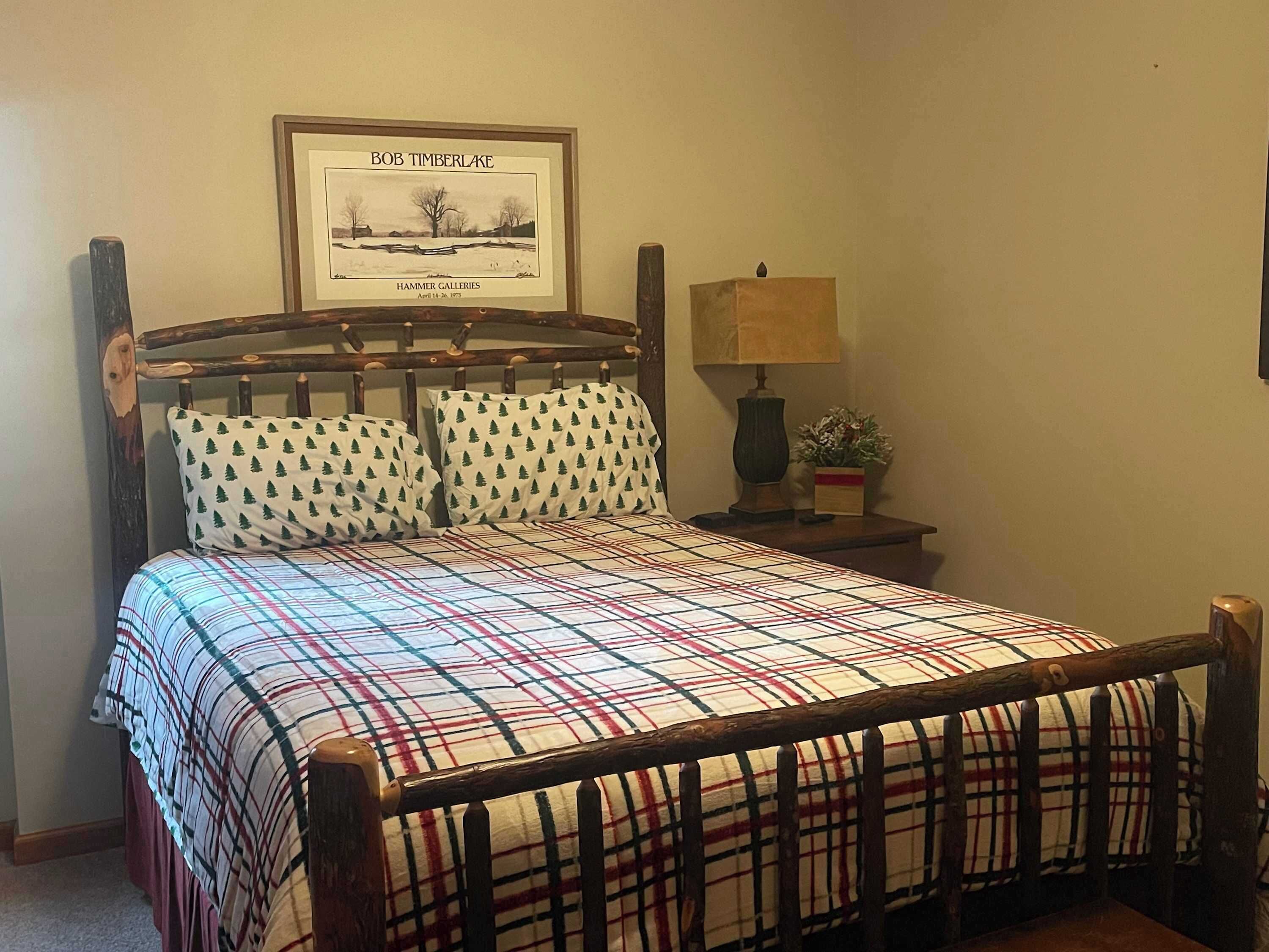 A bed with a plaid bedspread, a wooden headboard, and two matching pillows with a tree pattern. There is a framed picture above the bed and a small table with a lamp and a plant beside it.