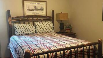 A bed with a plaid bedspread, a wooden headboard, and two matching pillows with a tree pattern. There is a framed picture above the bed and a small table with a lamp and a plant beside it.