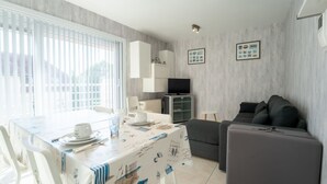 Un salon moderne avec un canapé gris, une table à manger recouverte d’une nappe blanche et un coin cuisine équipé d’un téléviseur et de placards.