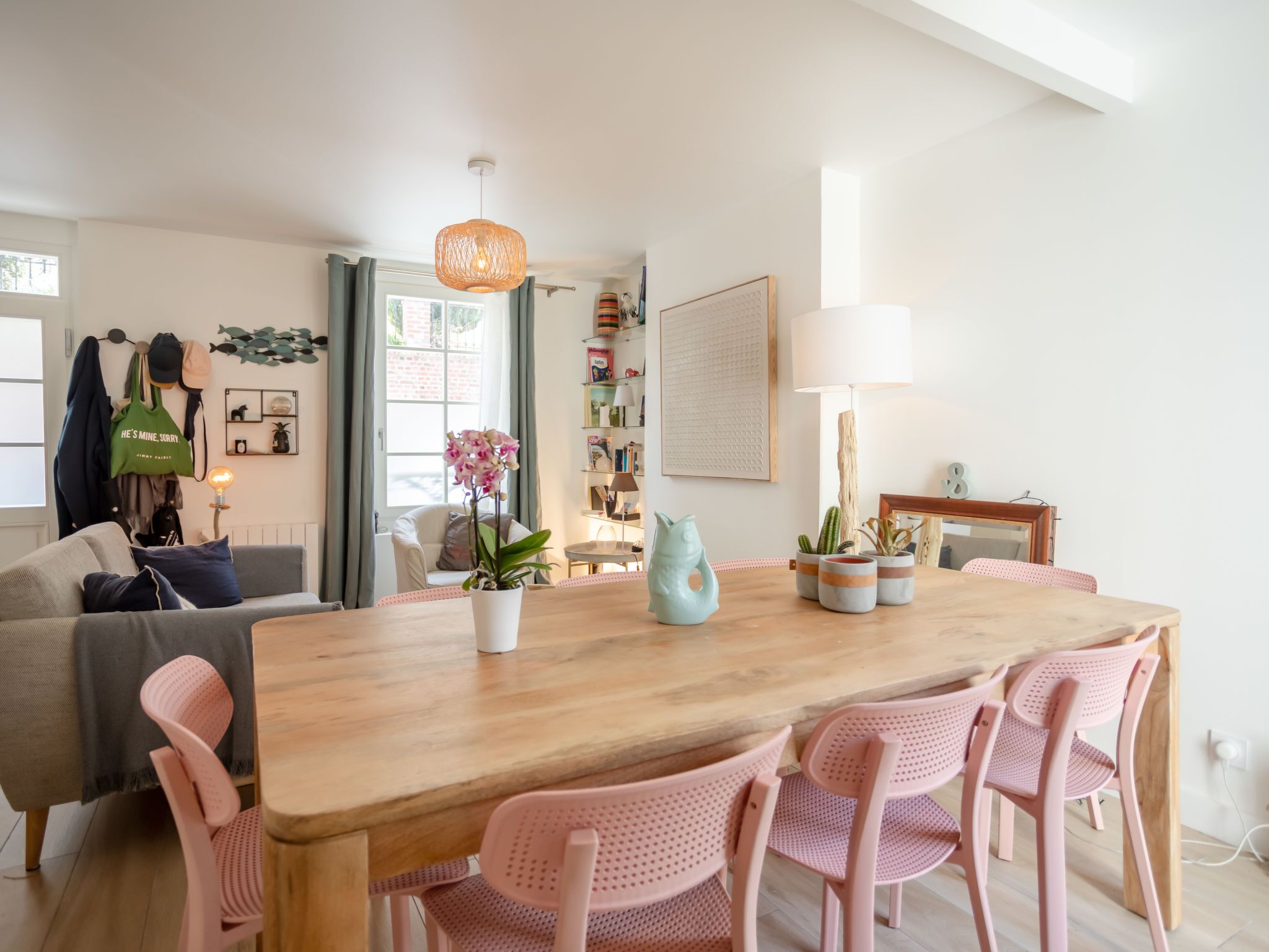 A modern living room with a wooden dining table, pink chairs, a white sofa, and a large window with curtains.