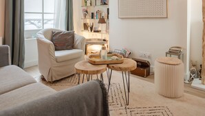 A cozy living room with a sofa, a coffee table, and a side table. There is a bookshelf with various books and decorative items. A window with curtains is visible, and a wall-mounted artwork is present. - La Trouvillaise by Interhome (Deauville-Trouville)