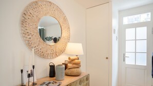 A modern interior with a round woven mirror, a wooden console table with a lamp, and a door with a window view. - La Trouvillaise by Interhome (Deauville-Trouville)