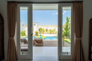 An open sliding door leading to a pool area with lounge chairs and a wooden deck. - Noel Peaceful 3BR Villa (Munggu)