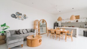 Een moderne woonkamer met een grijze bank, een houten salontafel, een eethoek met rieten stoelen, en een keuken met witte kastjes en een decoratieve spatwand.