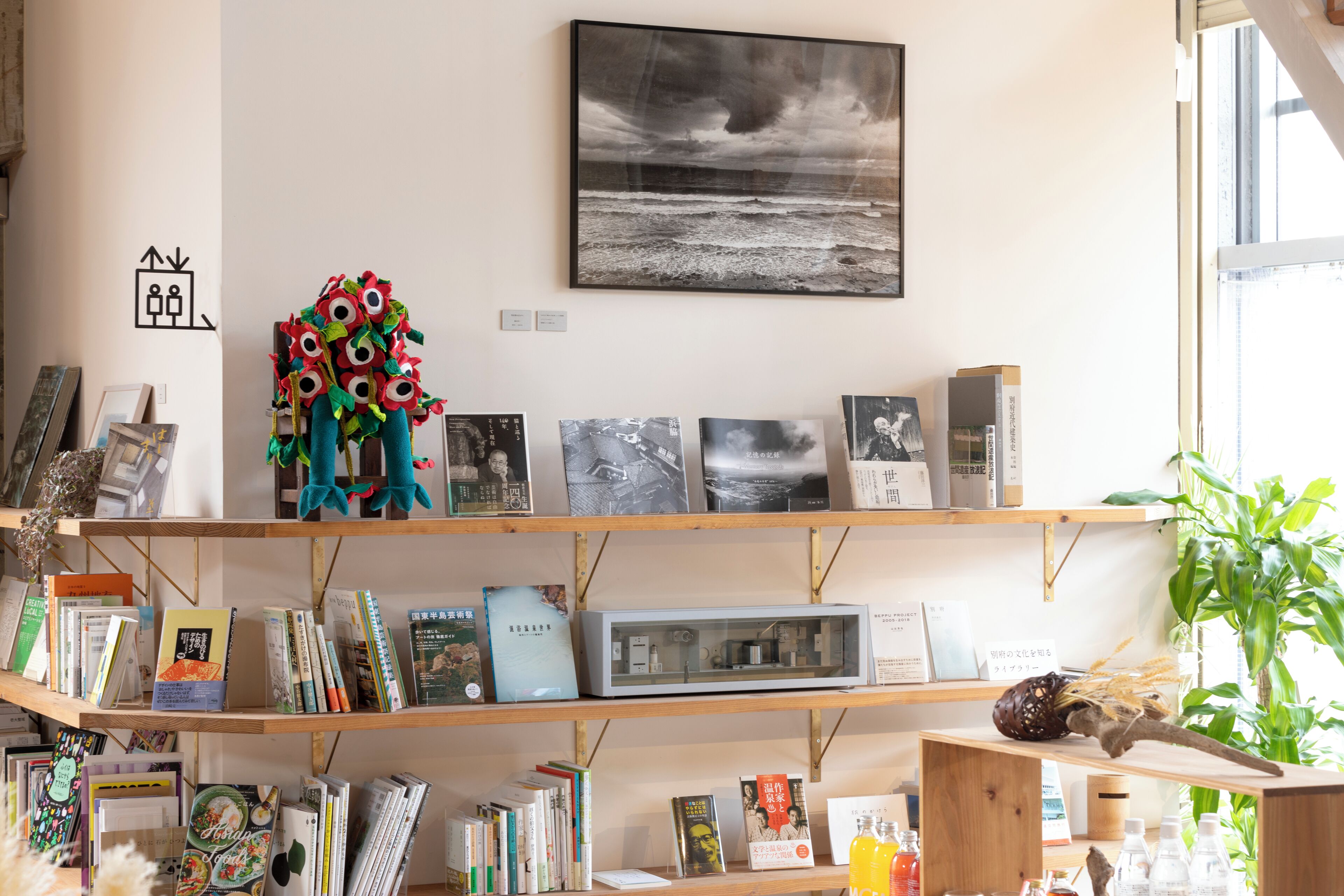 A room with a bookshelf filled with books and decorative items, a framed photograph on the wall, and a window with a view of the outdoors.