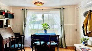 Ein gemütliches Zimmer mit Holztisch und -stühlen, einem Blumentopf und Blick ins Freie durch ein Fenster mit durchsichtigen Vorhängen.