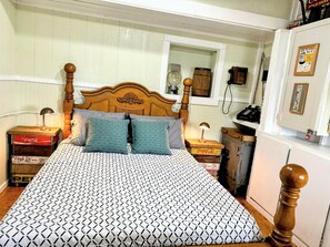 Een bed met een geborduurd sprei, houten hoofdbord en twee kussens. Twee nachtkastjes met lampen, een vintage telefoon en een koelkast met een foto in een lijst.