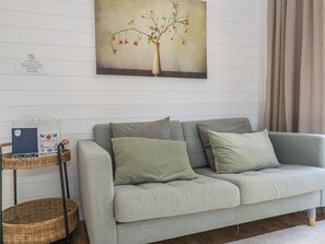 A grey sofa with two cushions, a wicker side table with a book, and a wall-mounted painting of a tree. - Villa sirius 56 by Interhome (Kemiönsaari)