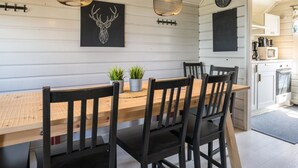 Un coin repas avec une table et des chaises en bois, deux lampes suspendues, une horloge murale et une cuisine visible à travers une porte ouverte.