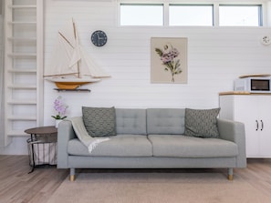 Una sala moderna con un sofá gris, una mesita redonda pequeña, un velero decorativo, un reloj, una obra de arte floral enmarcada y un microondas sobre un mueble.