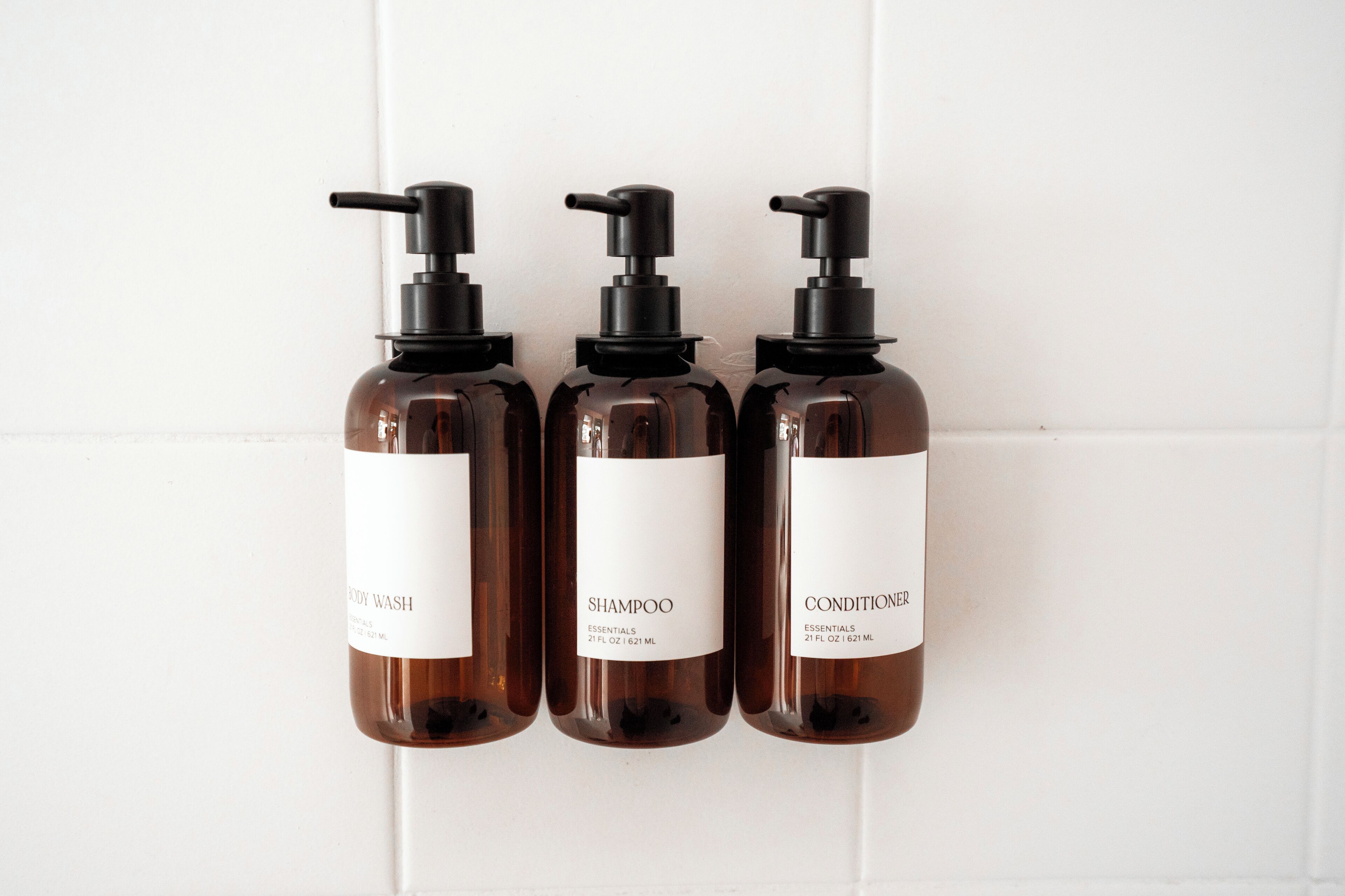 Three bottles of body wash, shampoo, and conditioner against a white tiled wall.