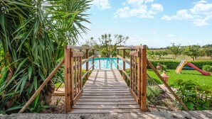 A wooden bridge leading to a swimming pool surrounded by greenery and a playground.