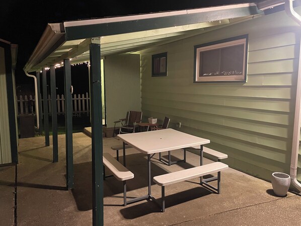 Un patio al aire libre con una mesa y bancos, una ventana pequeña y una valla blanca al fondo.