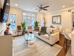 A modern living room with a sofa, coffee table, and chairs. There is a television mounted on the wall, a ceiling fan, and large windows.