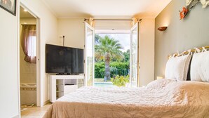 A bedroom with a bed, a TV, and a view of a swimming pool and palm trees.