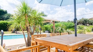 A wooden poolside table with an umbrella, surrounded by palm trees and a swimming pool.