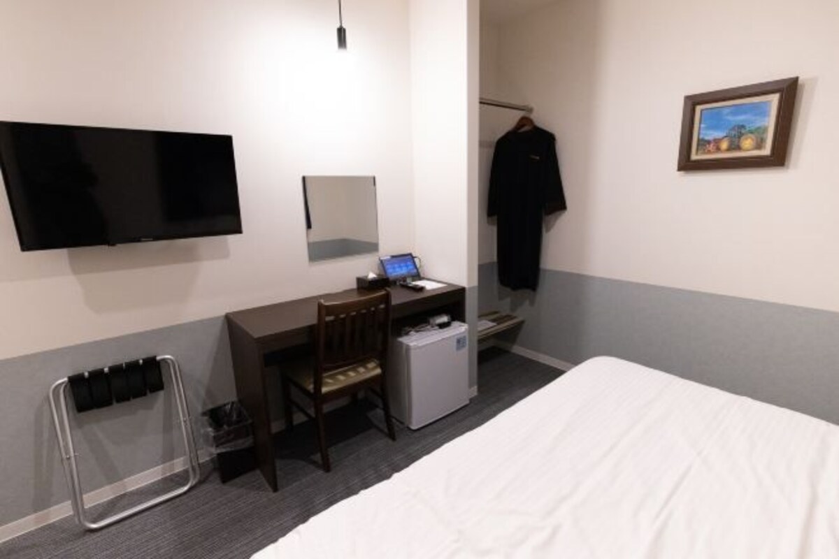 Habitación de hotel con cama, escritorio con computadora, televisión, perchero y refrigerador pequeño.