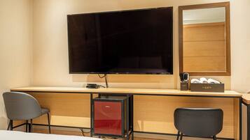 A hotel room with a bed, a desk with a flat-screen TV, a red mini-fridge, two chairs, and a wooden cabinet with toiletries.