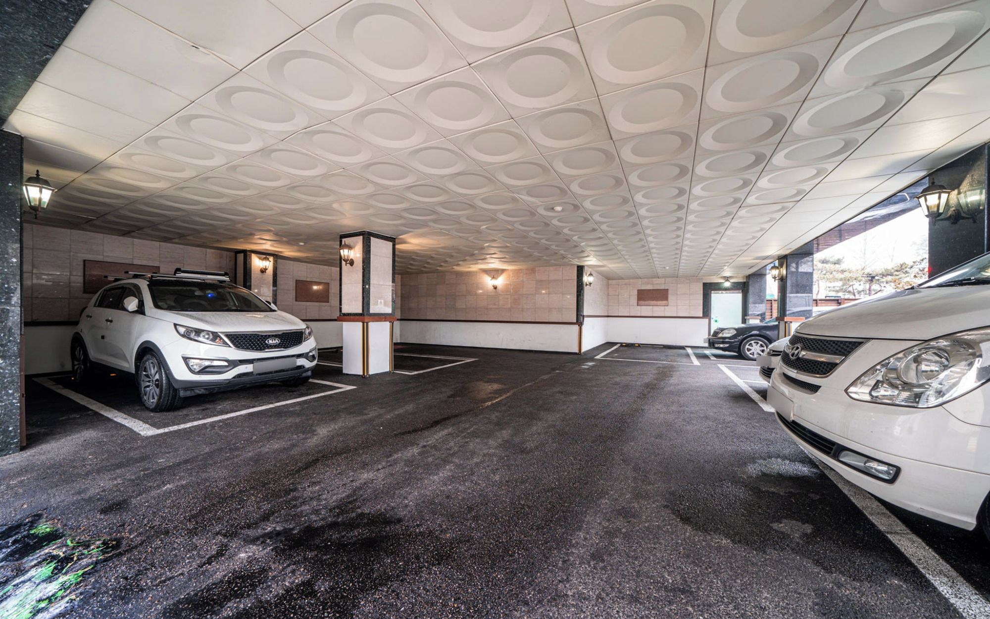 Un parcheggio multipiano con due auto, un SUV bianco e una berlina bianca, e un soffitto decorato con motivi circolari.