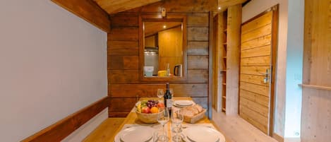 A wooden interior with a dining table set for two, a fruit bowl, wine glasses, and a basket of bread.