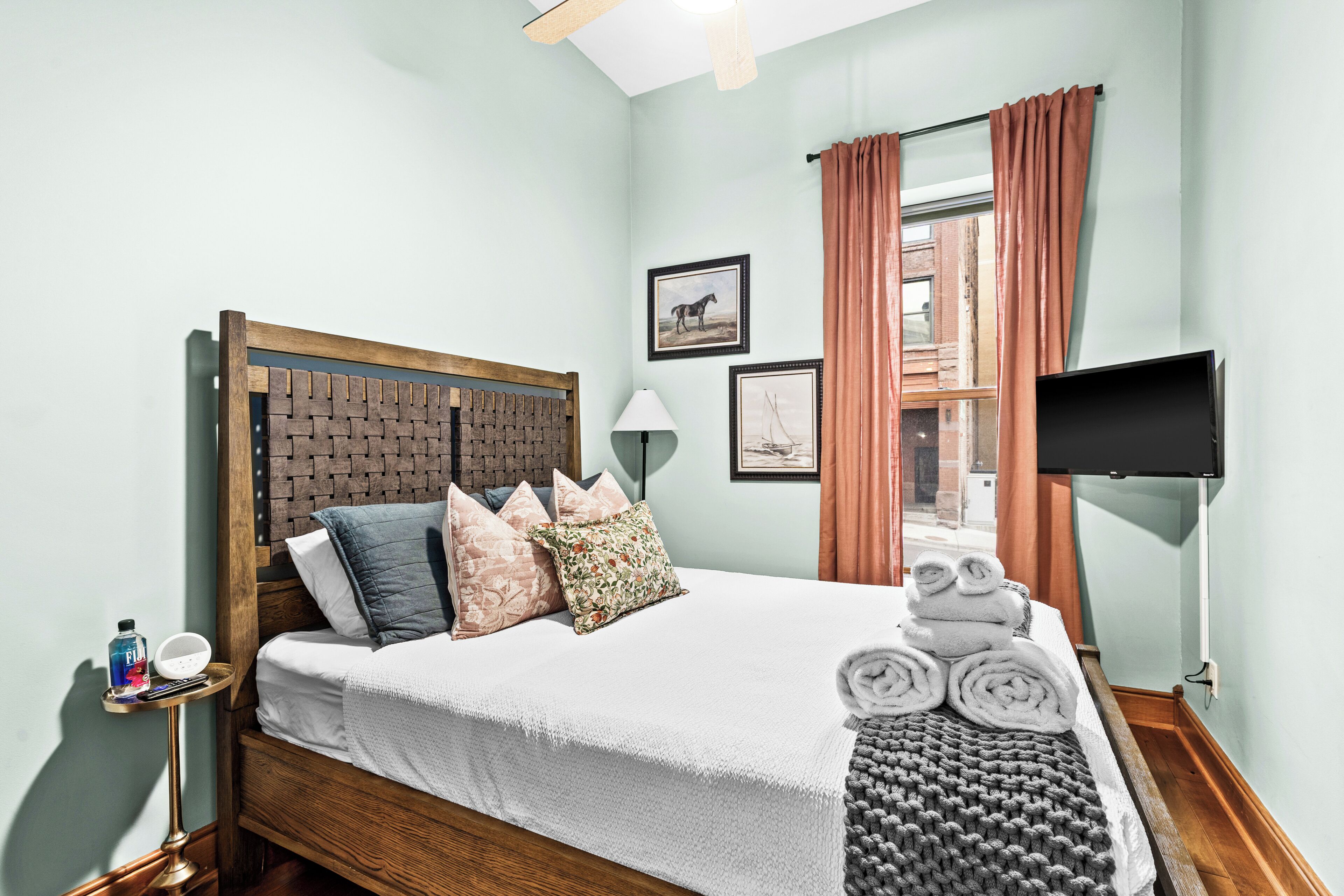 A neatly made bed with a wooden headboard, a bedside table with a lamp, and a window with curtains.