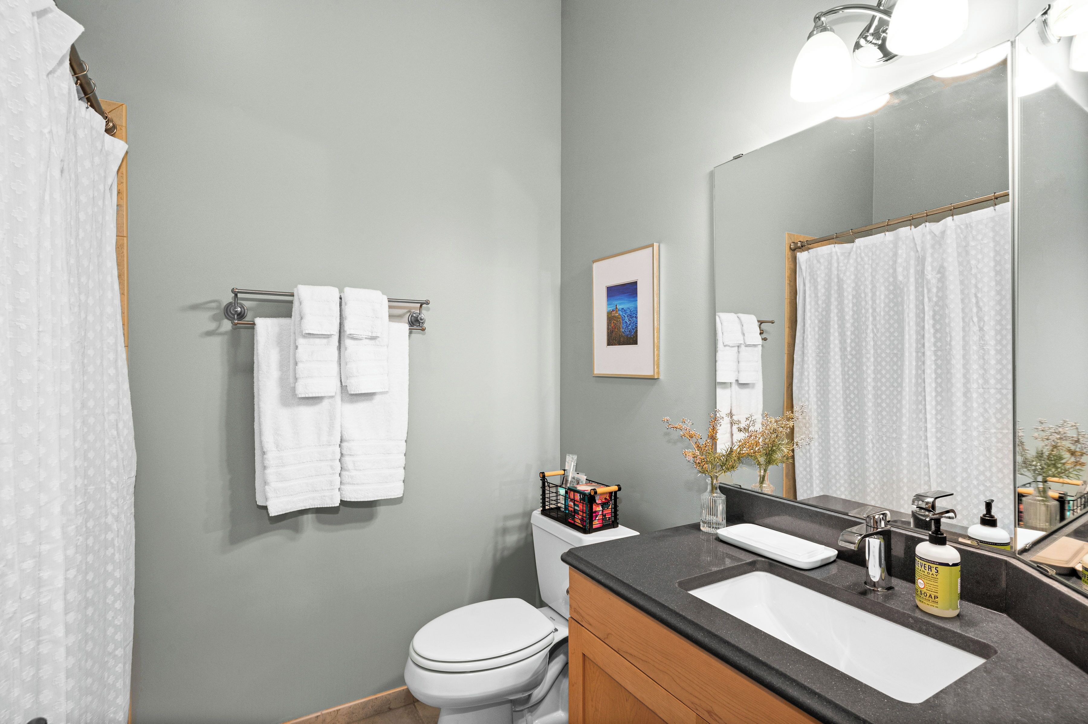 A bathroom with a toilet, sink, and mirror. There are towels hanging on a rack, a framed picture on the wall, and a basket of toiletries on the counter.
