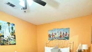A bedroom with a bed, a bench, a ceiling fan, a nightstand, and two paintings on the wall. - FLIP FLOP INN, 5MIN TO CRUISE AND BEACH (Galveston)