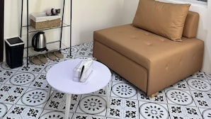 A room with a beige sofa, a white table, a black metal shelf with items, a patterned window, and a tiled floor. - Lanni House (Da Nang)