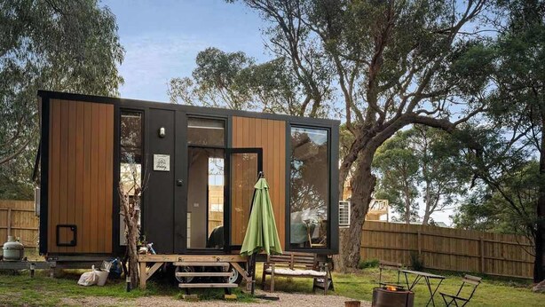 A modern, compact cabin with a wooden exterior and large windows, situated in a backyard with a fire pit and outdoor seating. - Metung Hideaway by Tiny Away (Metung)