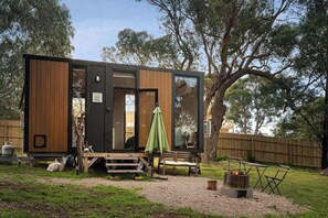 A modern, compact cabin with a wooden exterior and large windows, situated in a backyard with a fire pit and outdoor seating. - Metung Hideaway by Tiny Away (Metung)
