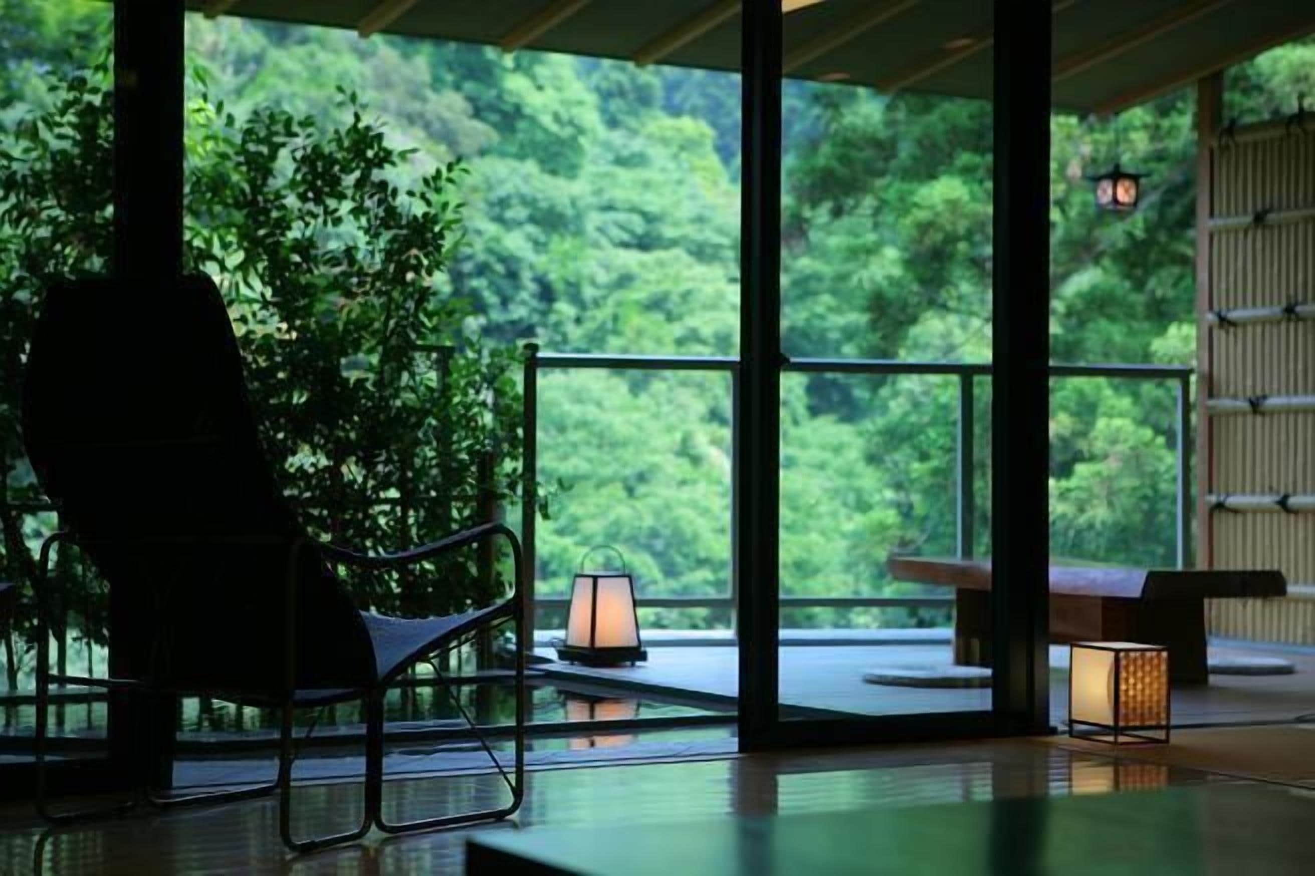 A room with a view of lush greenery, a chair, a table, and a lantern.