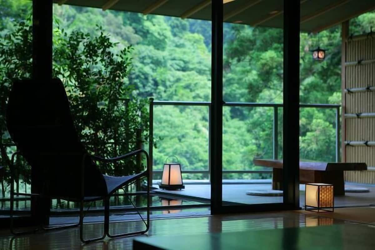 A room with a view of lush greenery, a chair, a table, and a lantern.