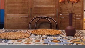 A dining area with a table set for two, a woven placemat, a vase with flowers, and a wooden partition with a circular light fixture above. - Warm and colourfull  studio in historical district of Chania (Chania)