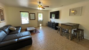 A living room with a leather couch, a coffee table, a dining table with stools, a ceiling fan, a flat-screen TV, and a door with a mirror. - Spacious Cottage in Rogers. Central: bike trails, restaurants, work, everything! (Rogers)