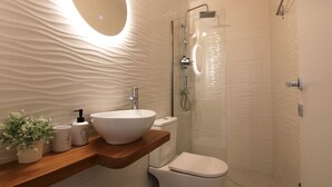 A modern bathroom with a large oval mirror, a vessel sink, a toilet, and a glass-enclosed shower.
