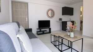 A modern living room with a grey sofa, a flat-screen TV, a small dining table, and a kitchen area with a black cabinet and a round mirror on the wall.