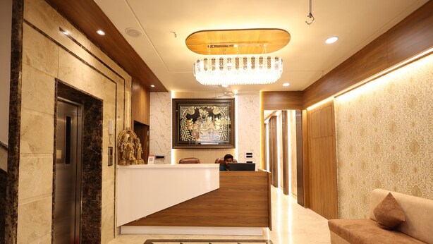 A hotel reception area with a marble floor, a wooden reception desk, a chandelier, and a framed artwork above the desk. - SHRI HARIDAS KUNJ (Mathura)