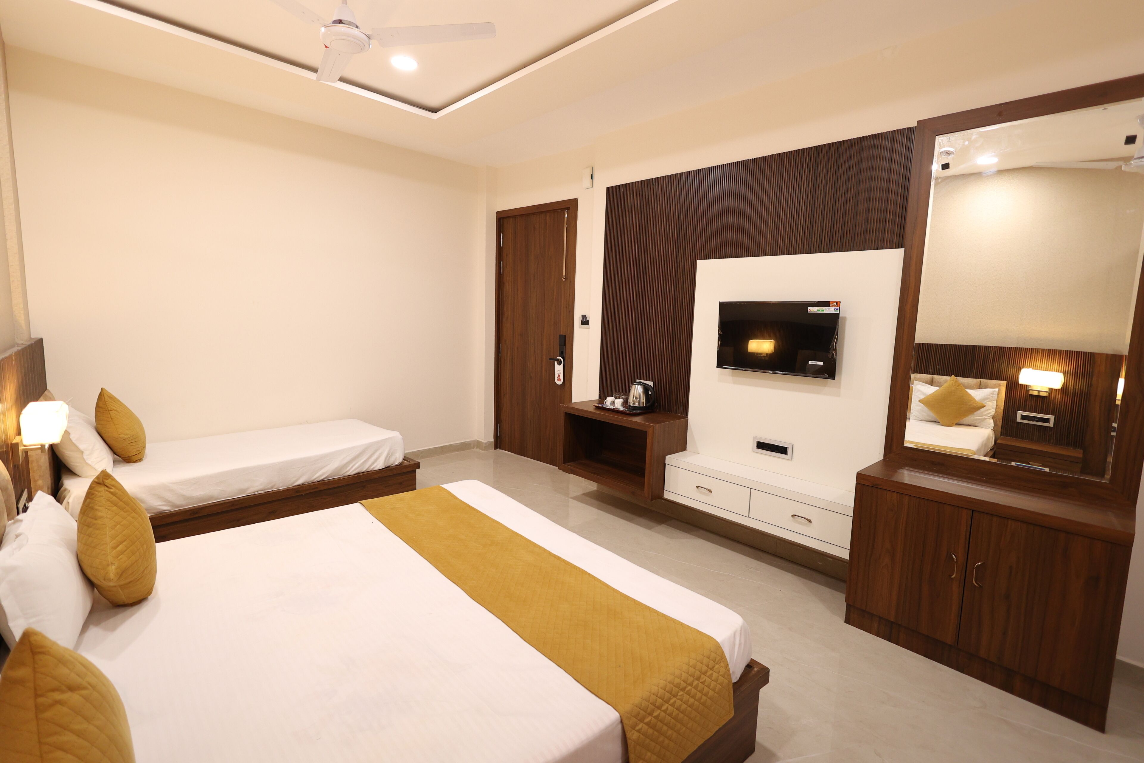 Une chambre d’hôtel avec deux lits simples, une télévision, un miroir, une armoire en bois et un ventilateur de plafond.