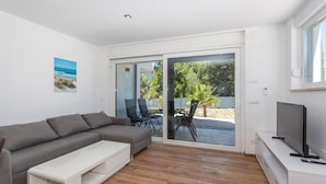A modern living room with a grey sofa, a white coffee table, and a TV on a white cabinet. There is a sliding glass door leading to an outdoor area with a patio table and chairs.